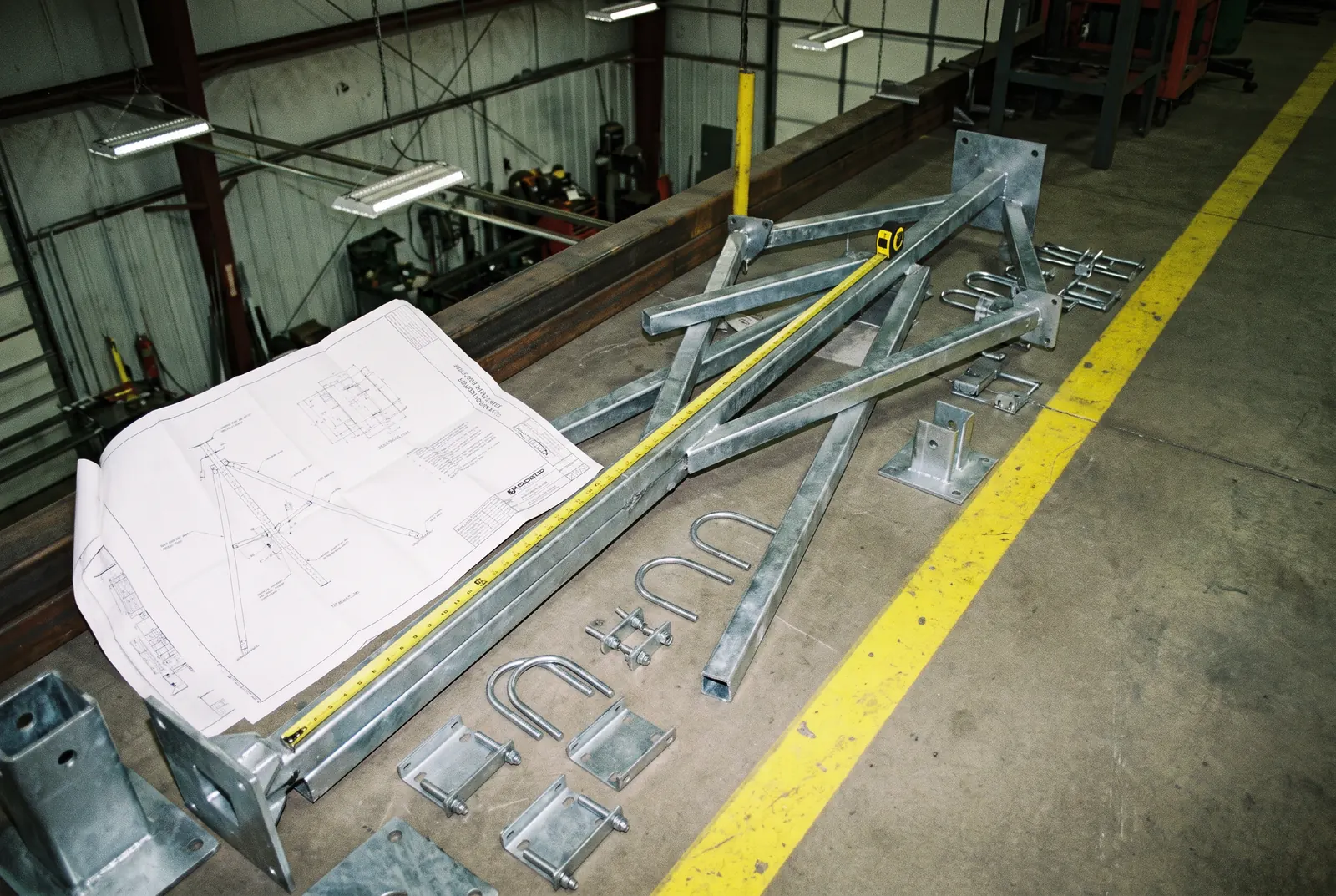 Support d'antenne en tripode galvanisé posé sur le plancher d'atelier à côté de son dessin, avec boulons en U et plaques de serrage